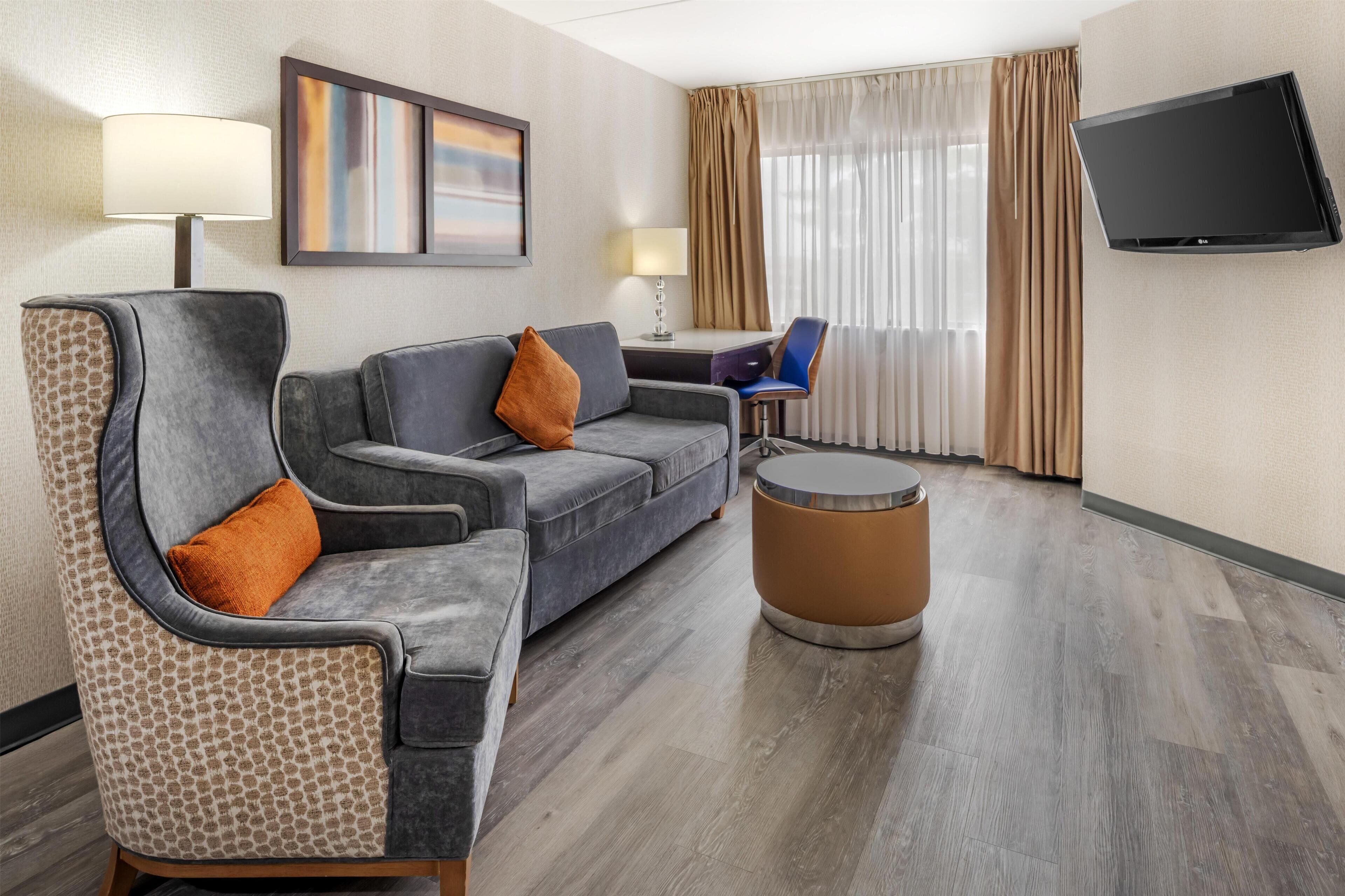 Een hotelkamer met een grijze bank, een gestreept fauteuil, een klein rond tafeltje, een aan de muur bevestigde televisie en een raam met gordijnen.