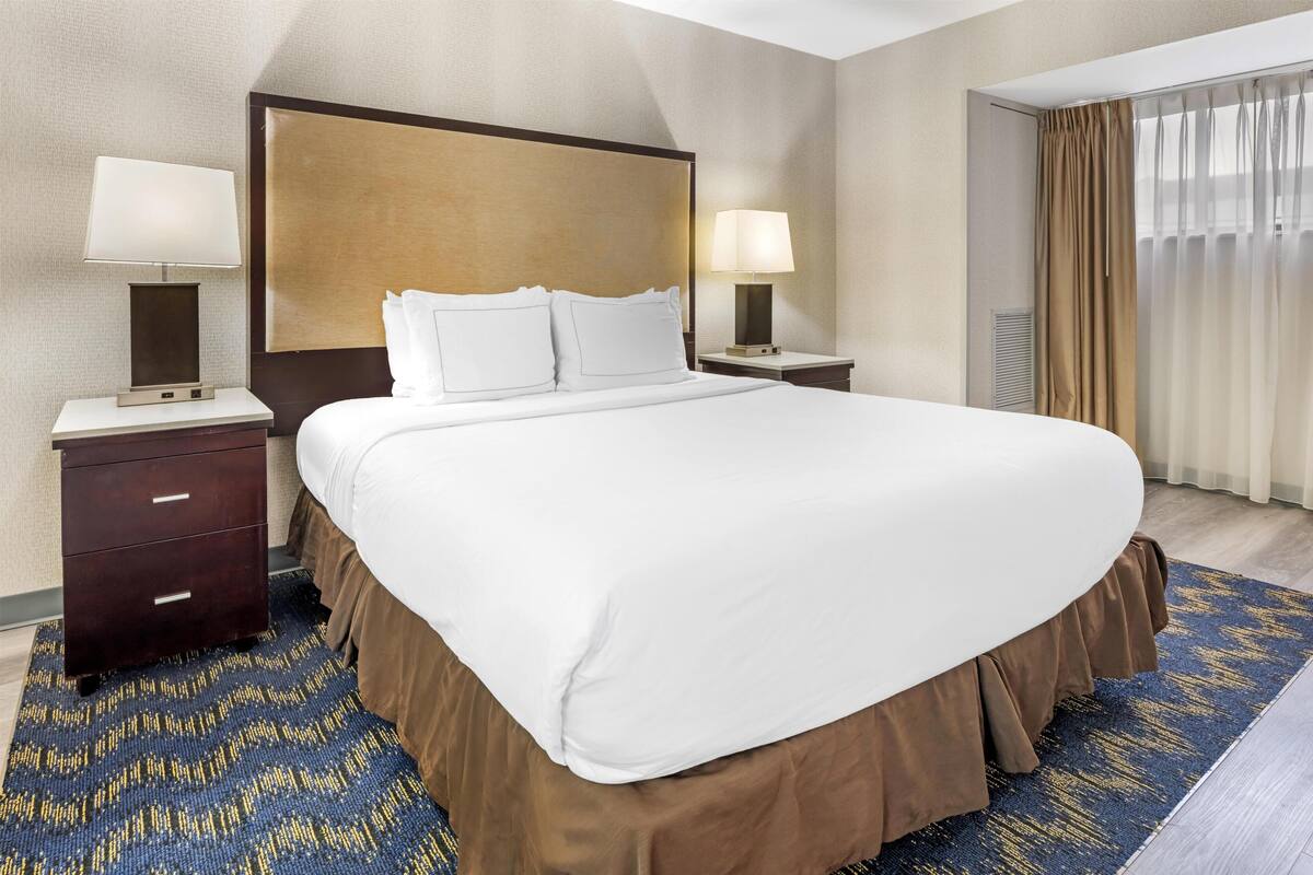 Ein Hotelzimmer mit einem großen Bett, zwei Nachttischen inklusive Lampen, einer Kopfteil und einem Fenster mit Vorhängen, sowie einem gemusterten Teppich.