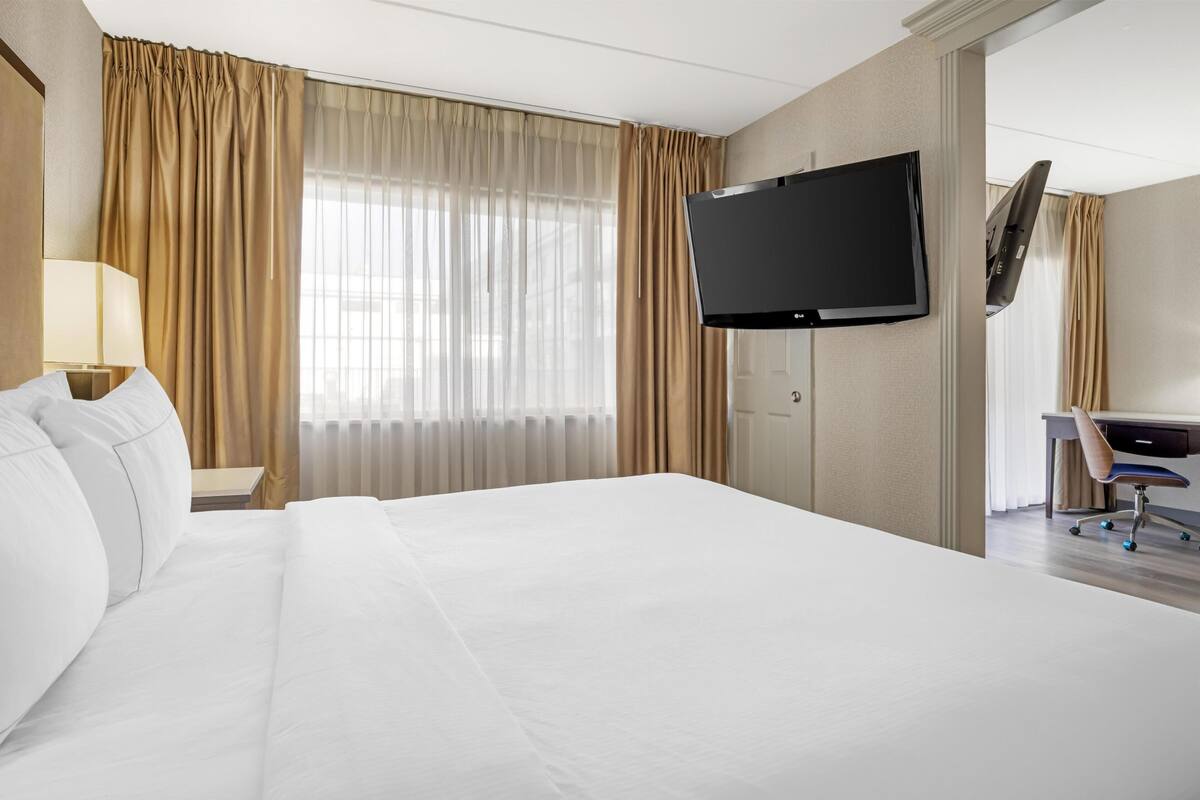 Ein Hotelzimmer mit einem großen Bett, einem flachbildfernseher an der Wand, einem Schreibtisch mit Stuhl und einem fenster mit Vorhängen.