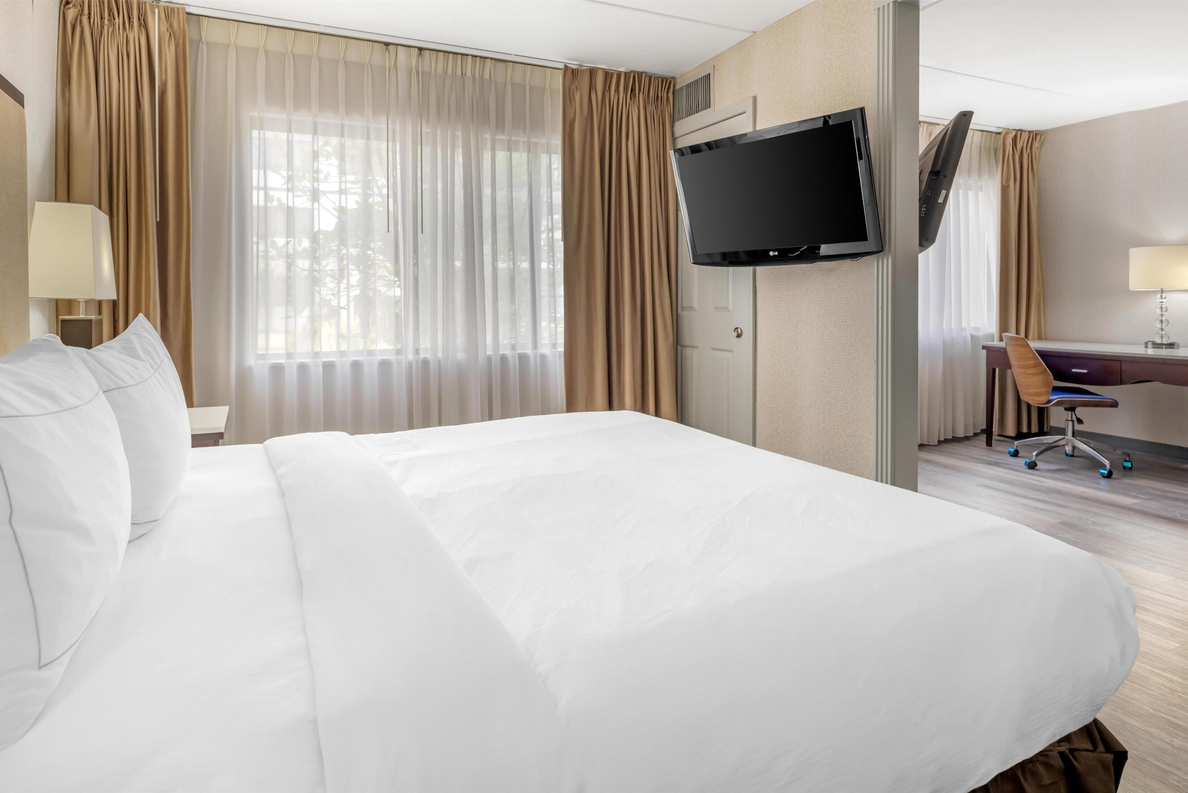 A hotel room with a large bed, a desk, a chair, a television mounted on the wall, and a window with curtains.