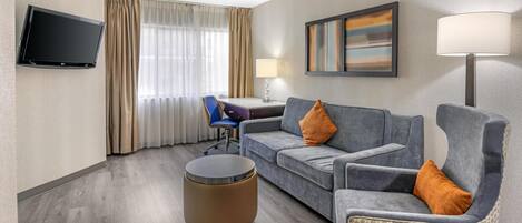 A hotel room with a flat-screen TV, a sofa, a chair, a desk, a lamp, and a painting on the wall.