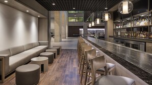 A modern bar area with a long counter, bar stools, and a seating area with a sofa and ottomans. - Hyatt Regency Schaumburg Chicago (Schaumburg)