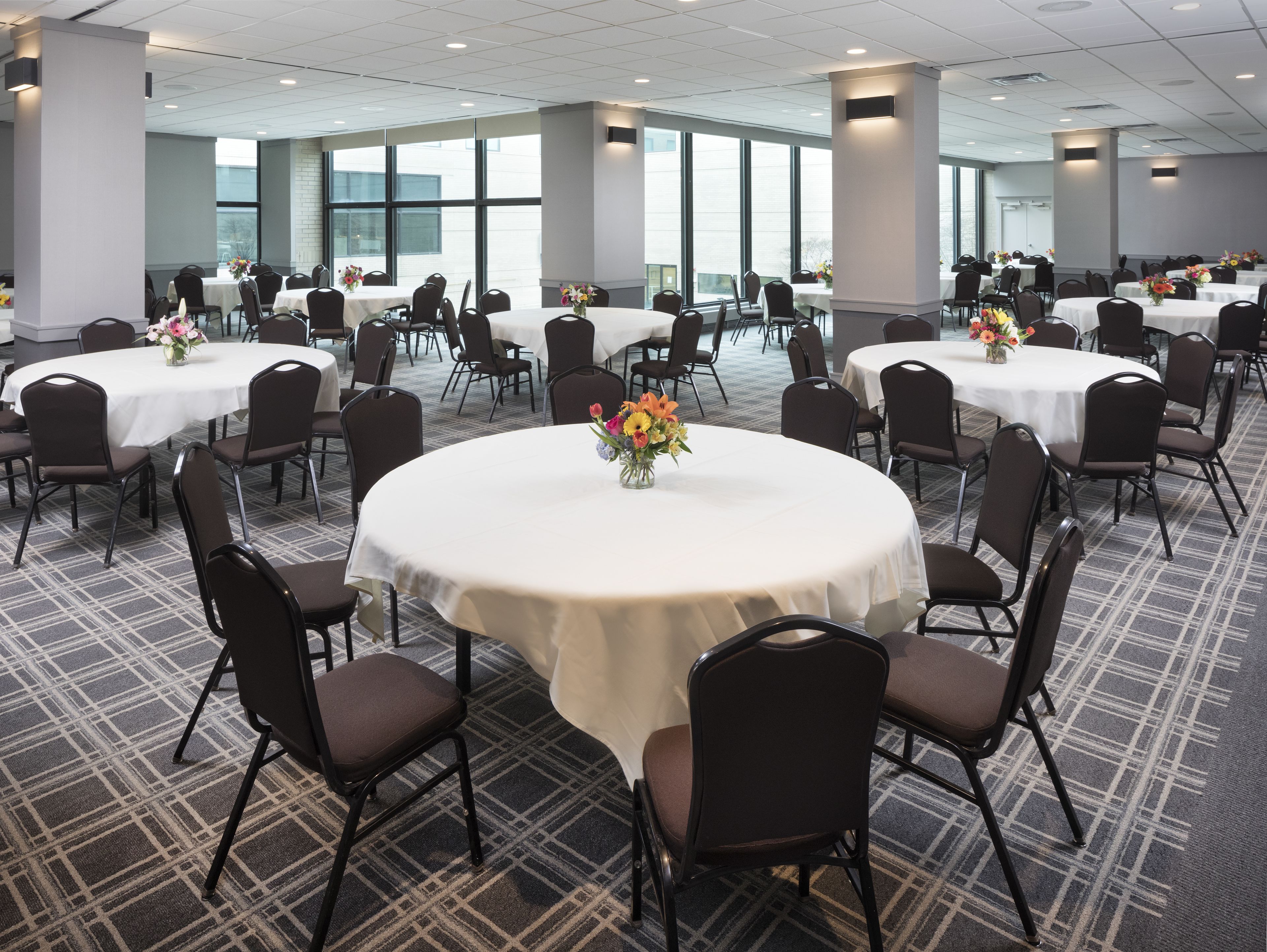 Une salle de conférence dotée de plusieurs tables rondes, chacune entourée de chaises, et de grandes fenêtres laissant entrer la lumière naturelle.