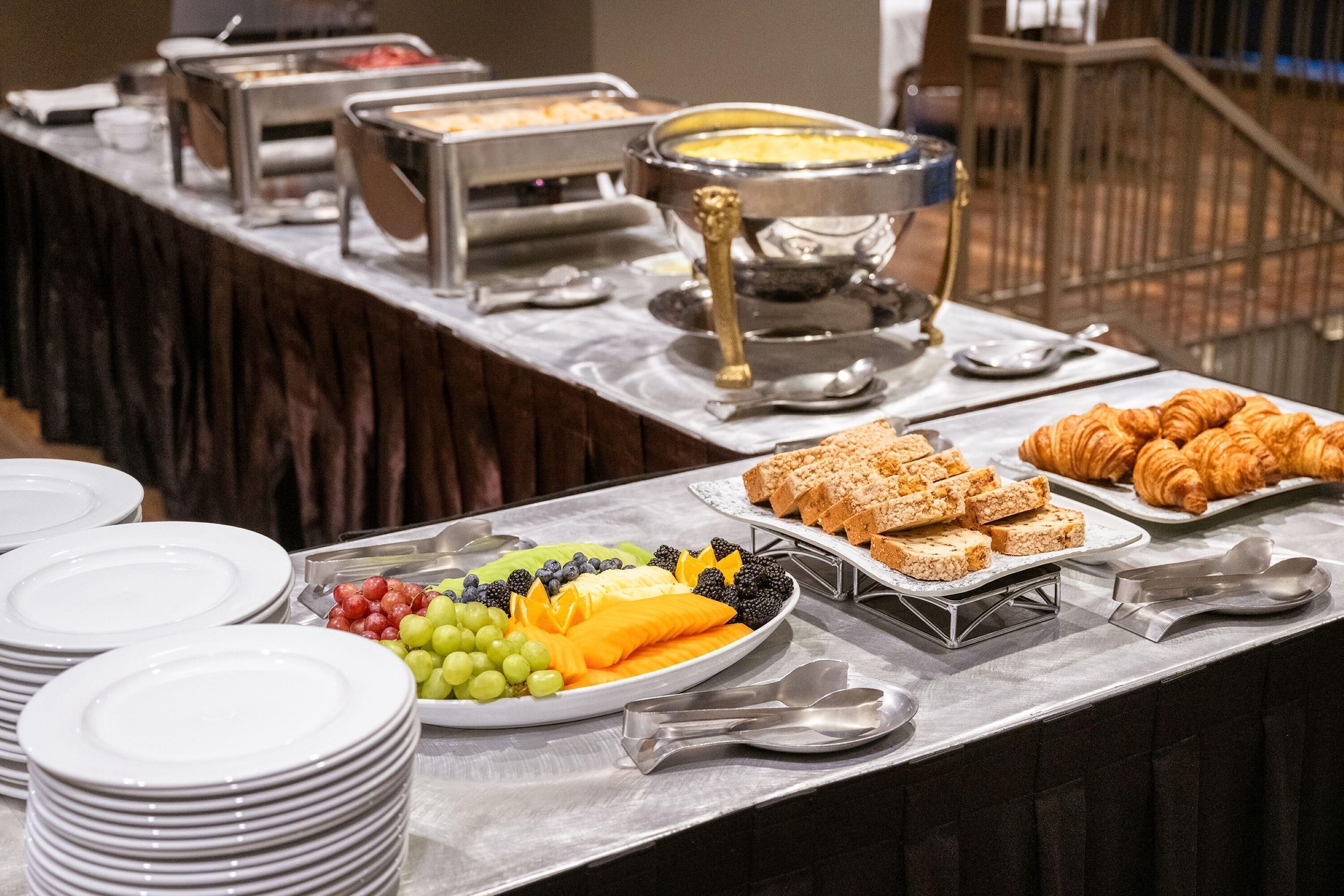 A buffet table with various food items including fruits, bread, and pastries.
