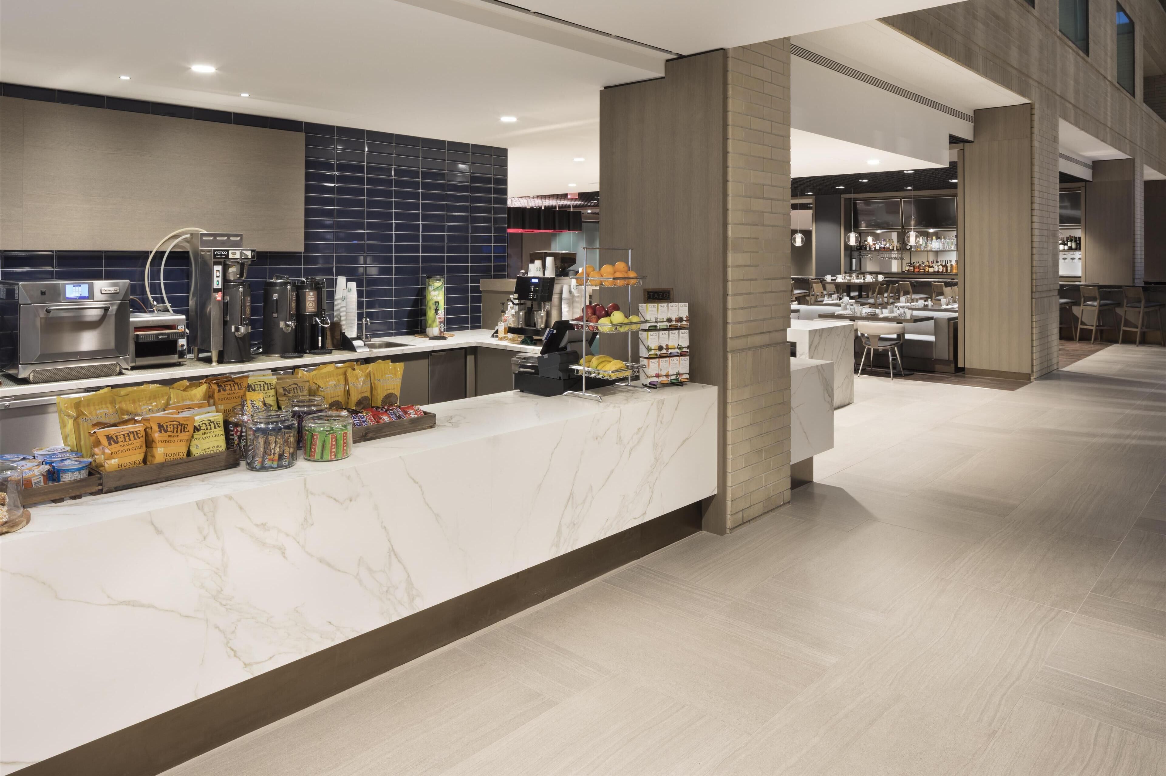 A modern hotel lobby with a marble reception desk, coffee machines, and a fruit display.