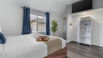 A modern hotel room with a large bed, a flat-screen TV mounted on the wall, a small plant, and a white shelving unit.