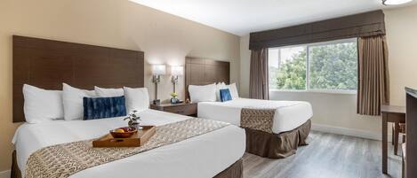A hotel room with two beds, a wooden headboard, a bedside table with lamps, a window with curtains, and a view of greenery outside.