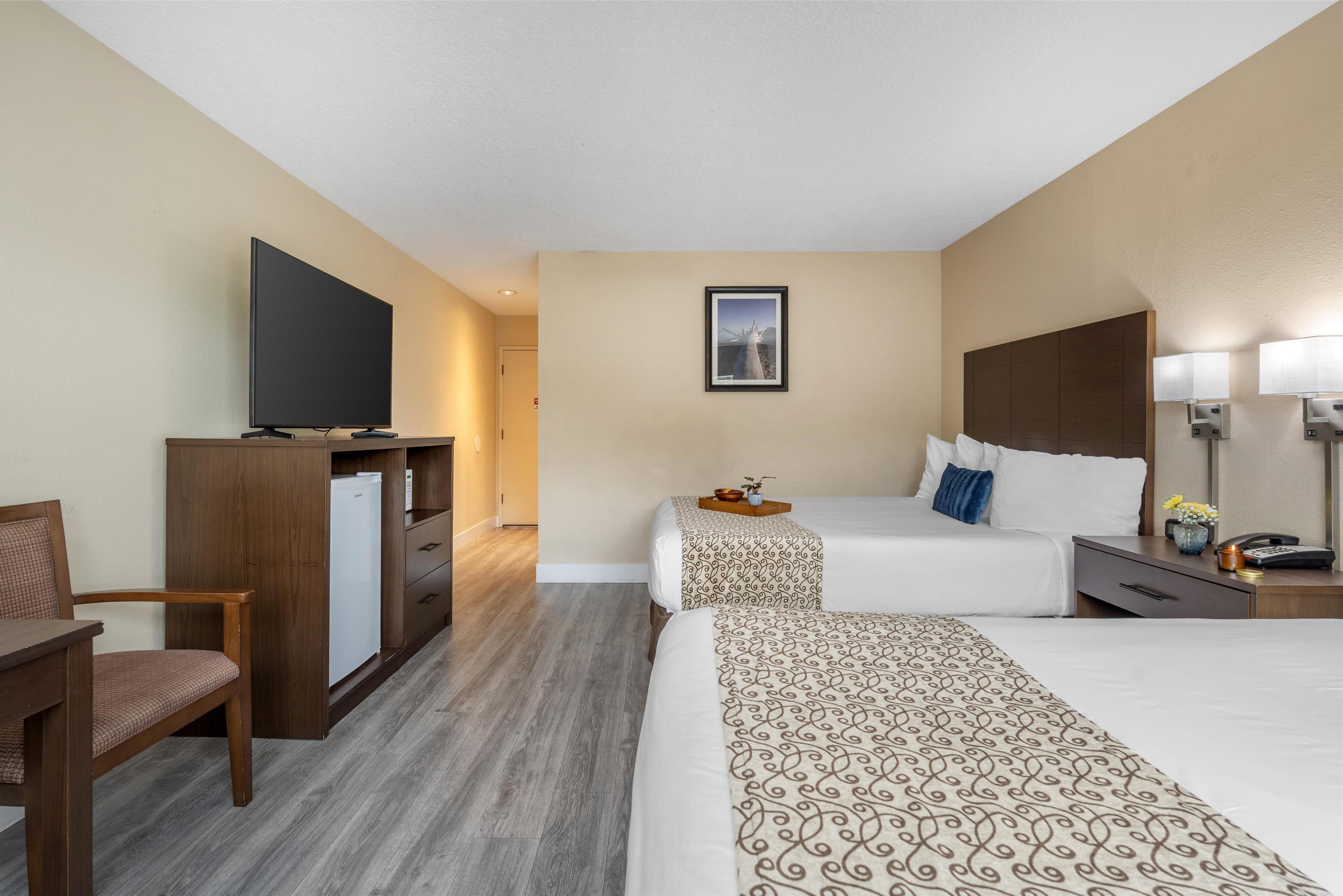 A hotel room with two beds, a desk, a TV, and a refrigerator.