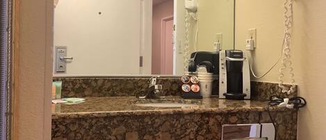 A bathroom with a granite countertop, a mirror, a hairdryer, a coffee maker, and a towel dispenser.
