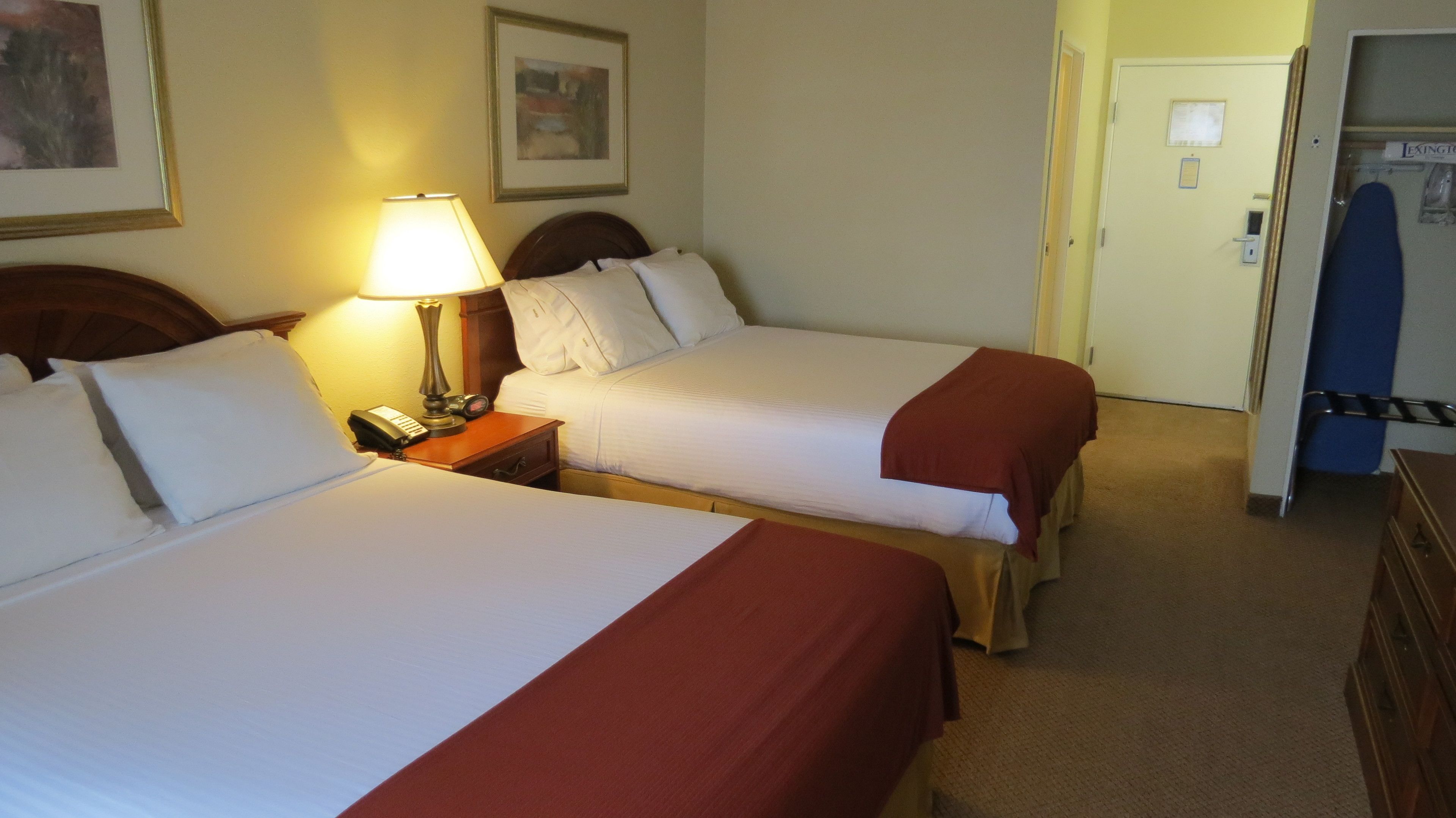 Een hotelkamer met twee bedden, een nachtkastje met een lamp, een telefoon en een deur die naar een andere kamer leidt.