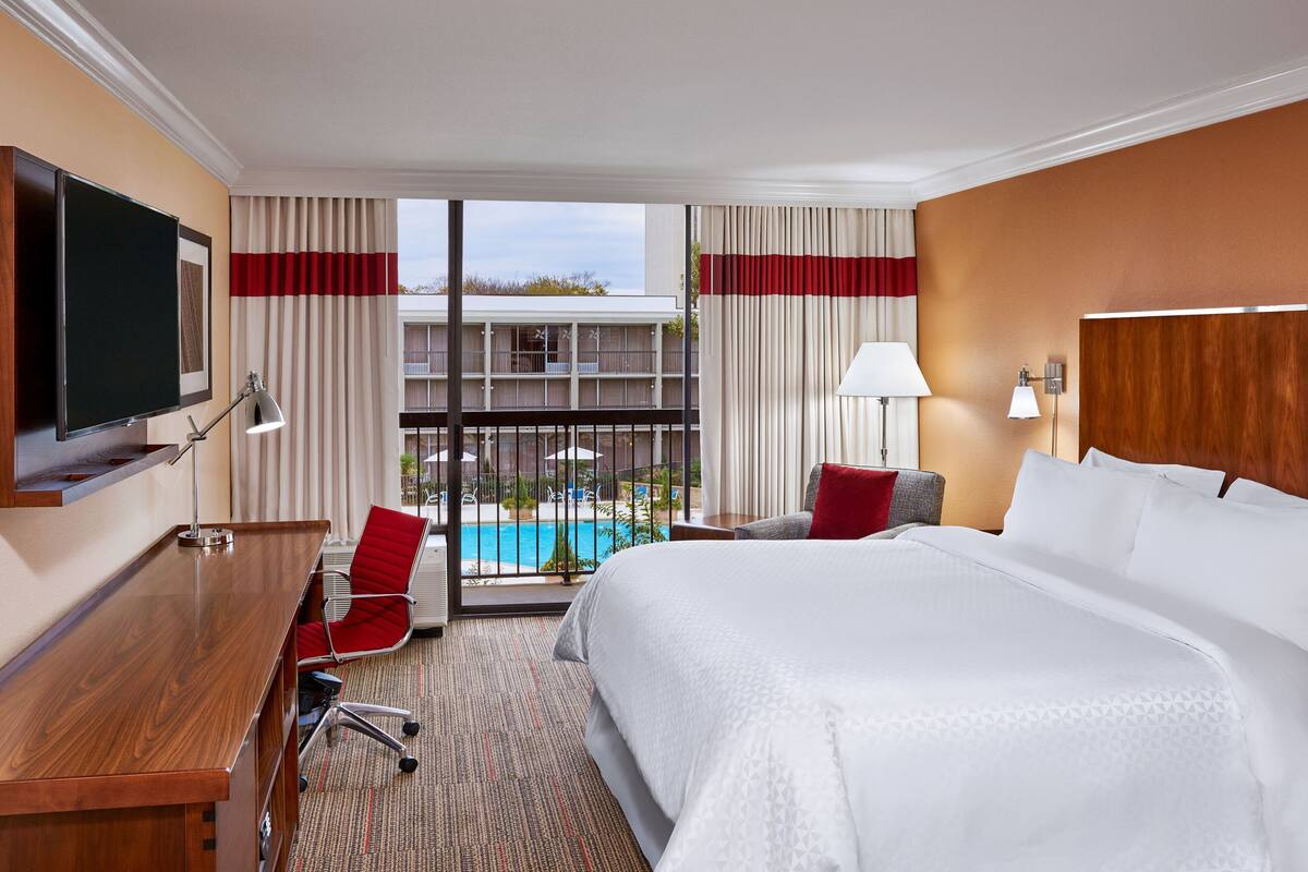 A hotel room with a large bed, a desk, a chair, a TV, a pool view, and a balcony.