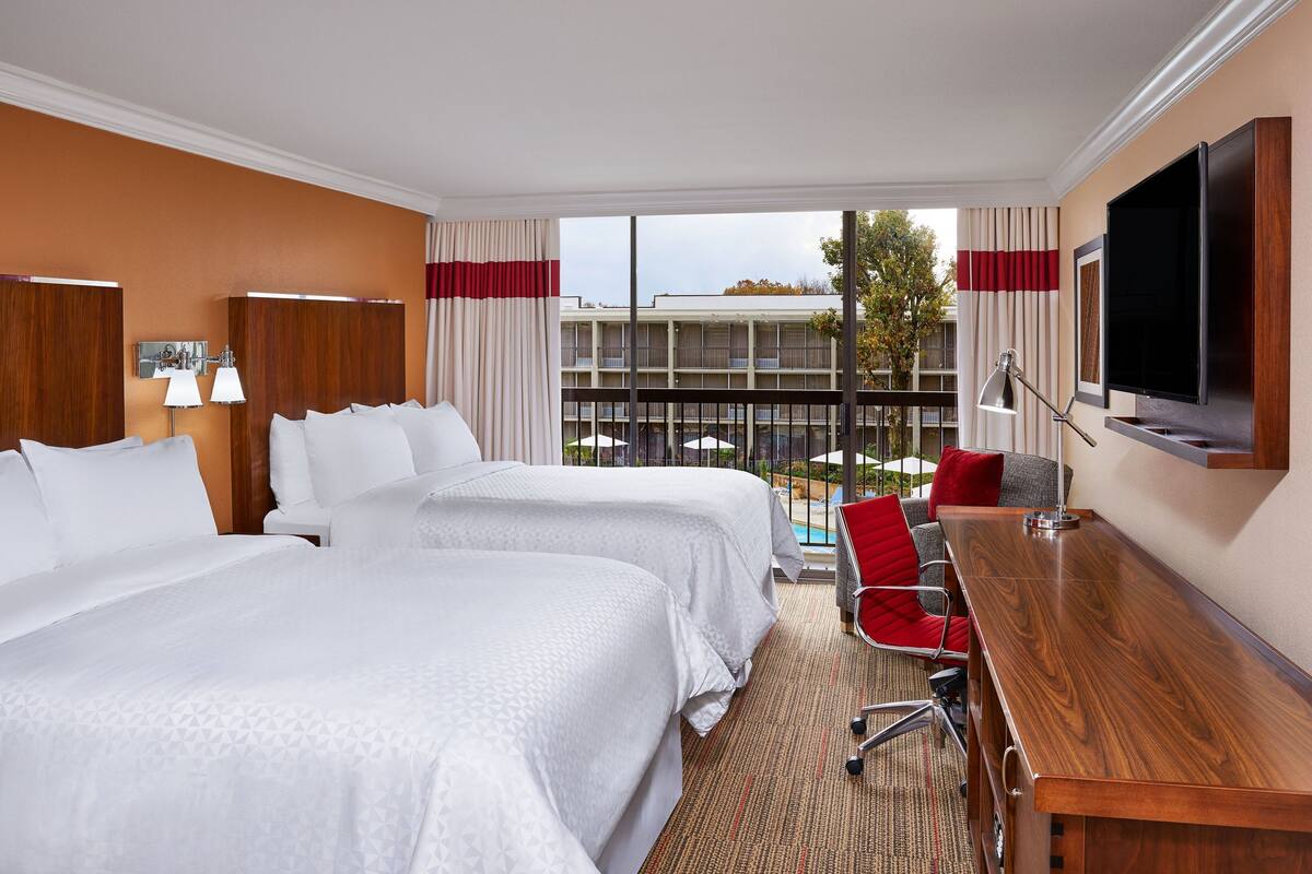 A hotel room with two beds, a desk, a TV, and a balcony with a pool view.