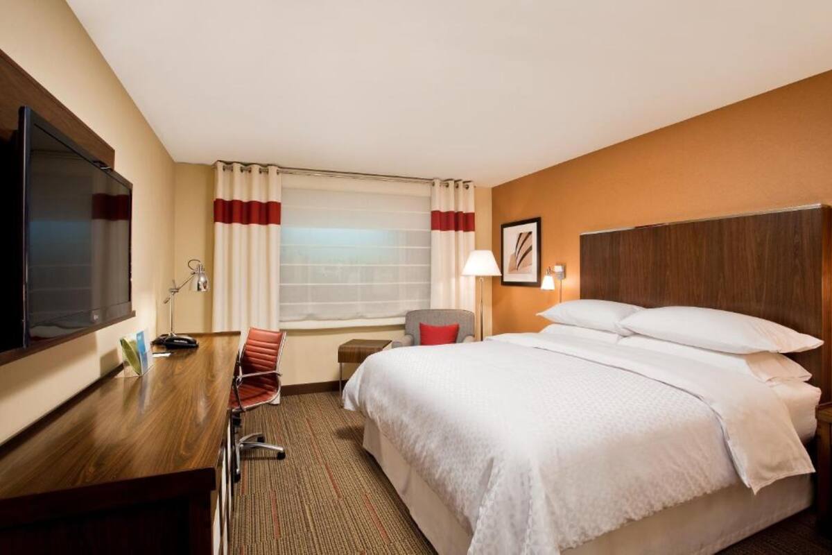 A hotel room with a bed, a desk, a chair, a TV, a lamp, and a window with curtains.