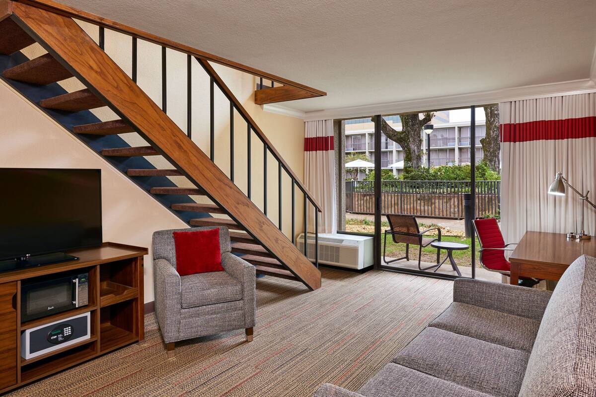 A living room with a wooden staircase, a flat-screen TV, a grey armchair, a red pillow, and a sofa.