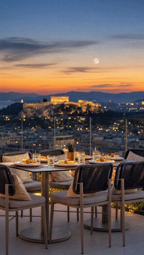 An outdoor dining area with tables and chairs, a view of a cityscape, and a distant illuminated building. - St. George Lycabettus Lifestyle Hotel (Athens)