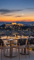 An outdoor dining area with tables and chairs, a view of a cityscape, and a distant illuminated building.