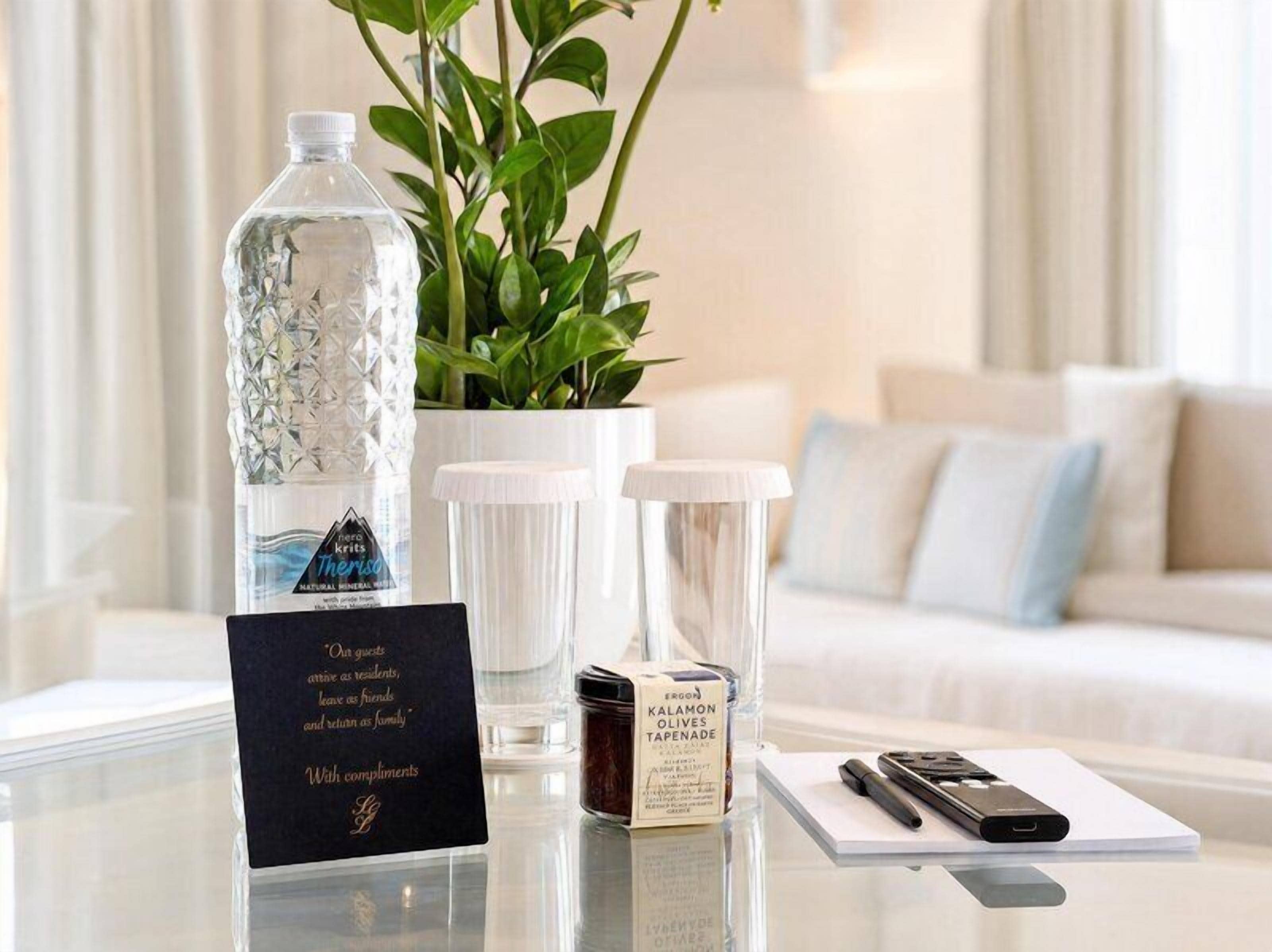 Habitación de hotel con una mesa de vidrio que muestra una botella de agua, una planta, una vela, una caja de artículos de tocador y una libreta.