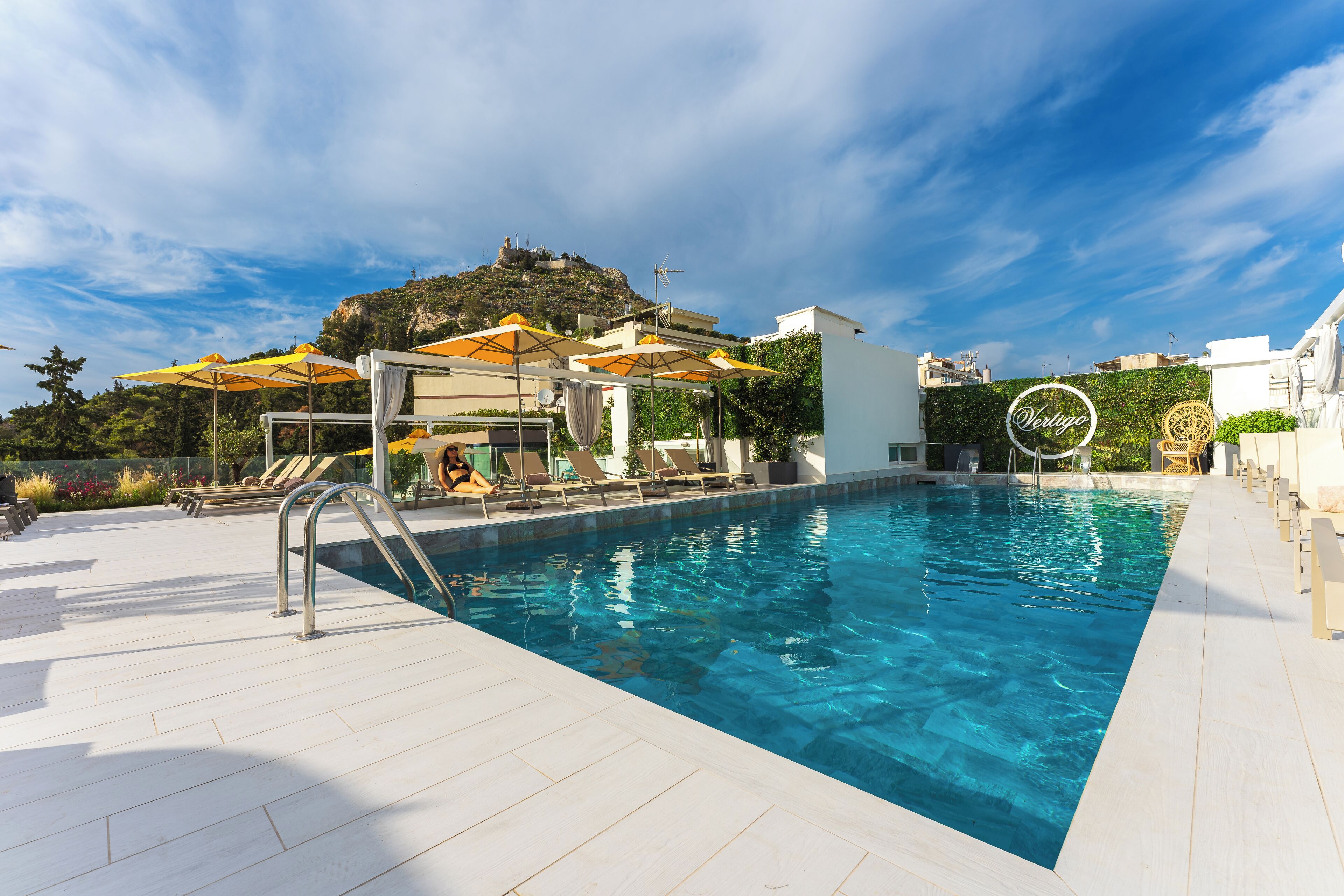 A swimming pool with sun loungers and umbrellas, a white building, and a hill with a statue in the background.