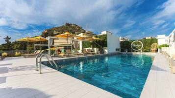 A swimming pool with sun loungers and umbrellas, a white building, and a hill with a statue in the background.