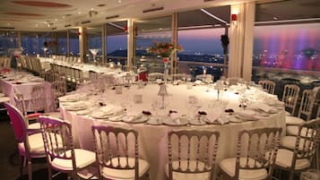 Un salón de banquetes con mesas redondas dispuestas para un evento formal, amplias ventanas con vistas a la ciudad y un diseño interior moderno.