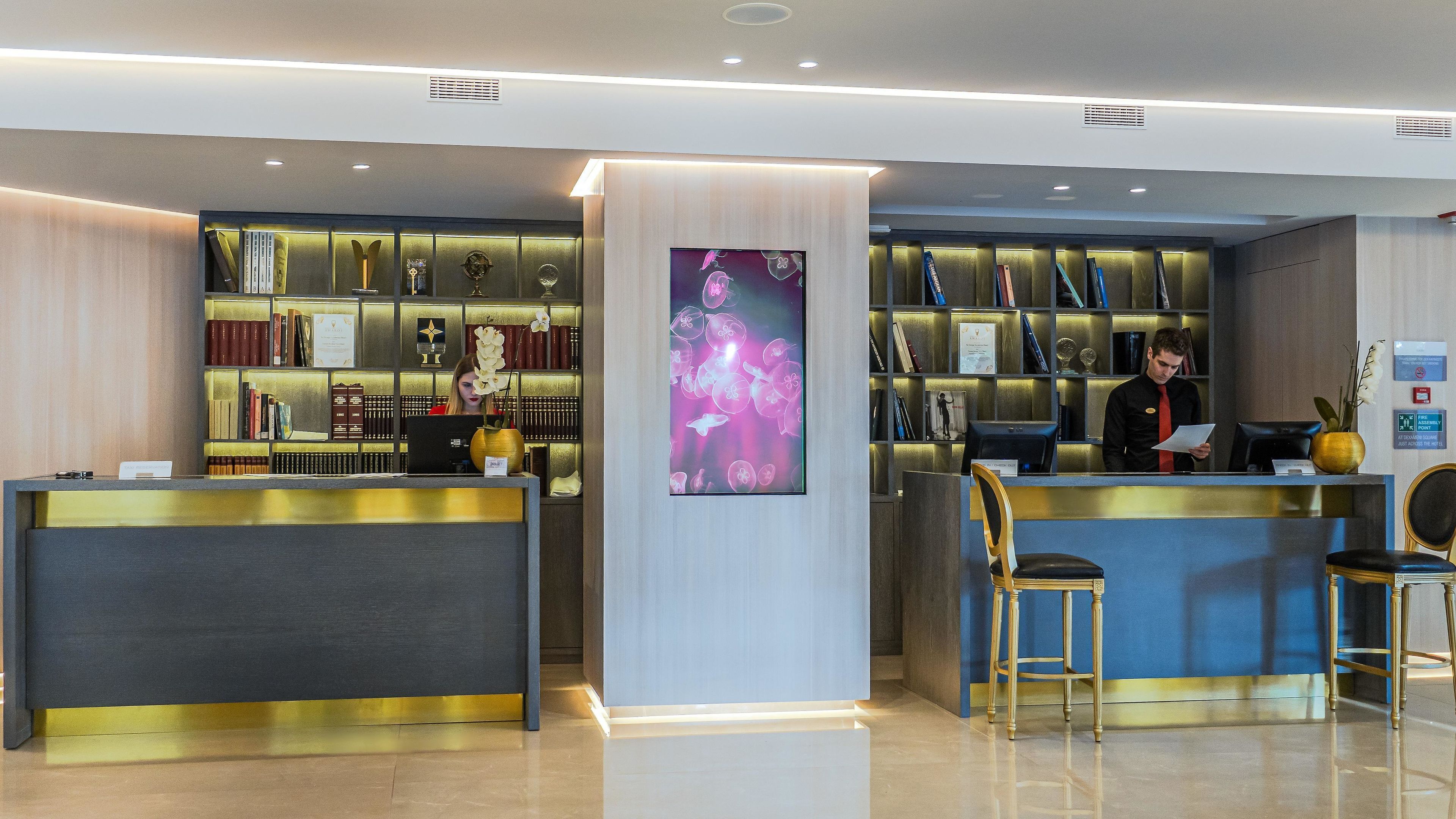A reception area with a modern design, featuring a reception desk, a bookshelf with various items, and a digital screen displaying an image.