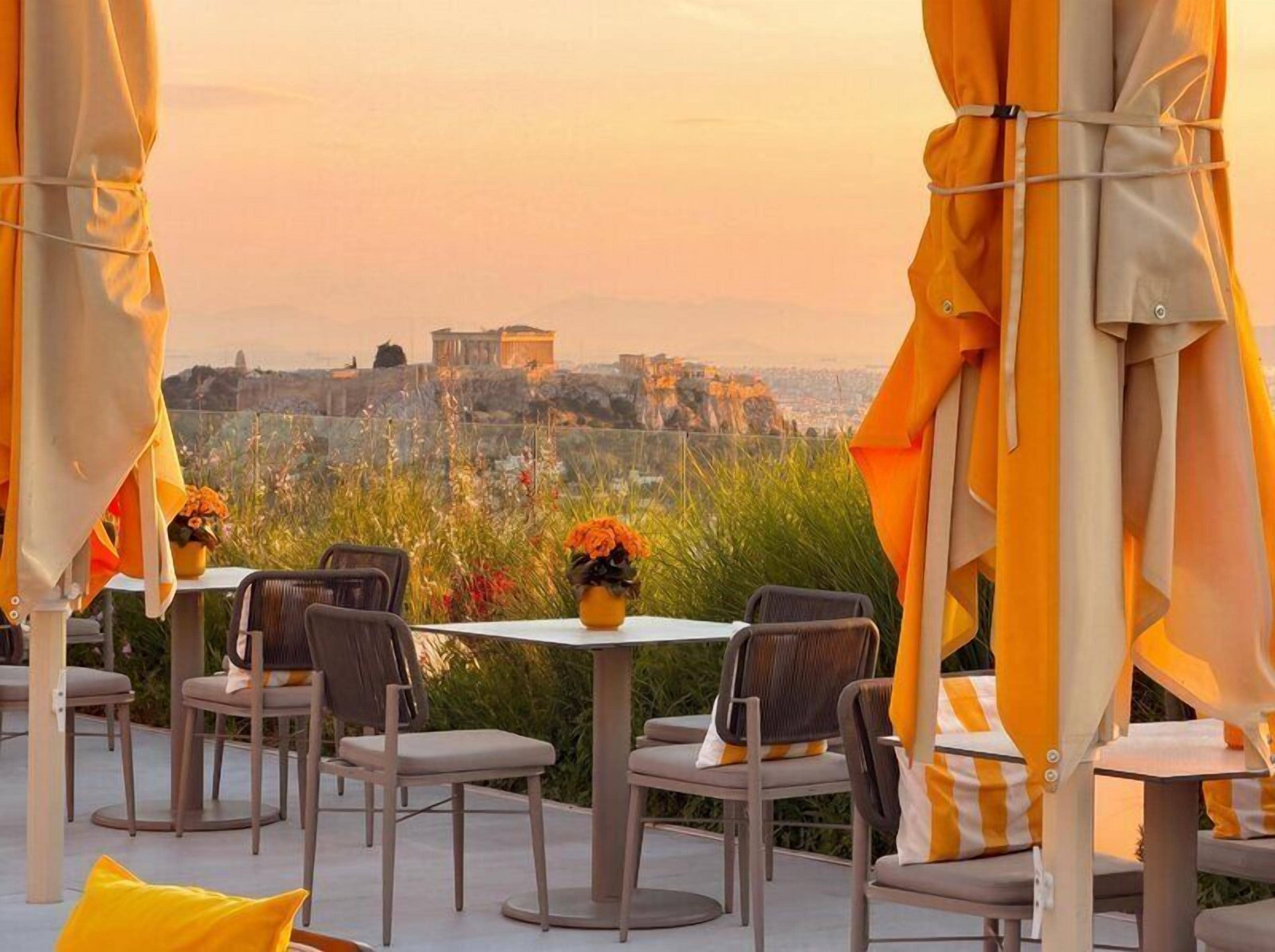 An outdoor seating area with tables and chairs, a view of a distant building, and a sunset.