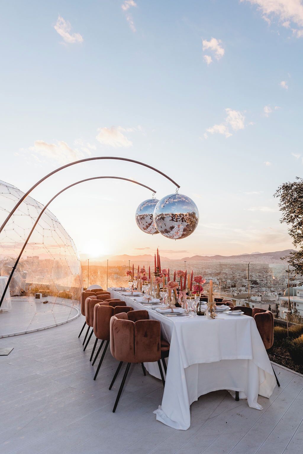 An outdoor dining setup with a clear view of the cityscape, featuring a long table with white tablecloths and brown chairs, and two disco balls above.