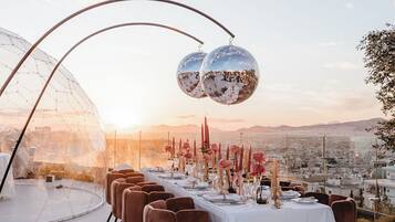 An outdoor dining setup with a clear view of the cityscape, featuring a long table with white tablecloths and brown chairs, and two disco balls above.