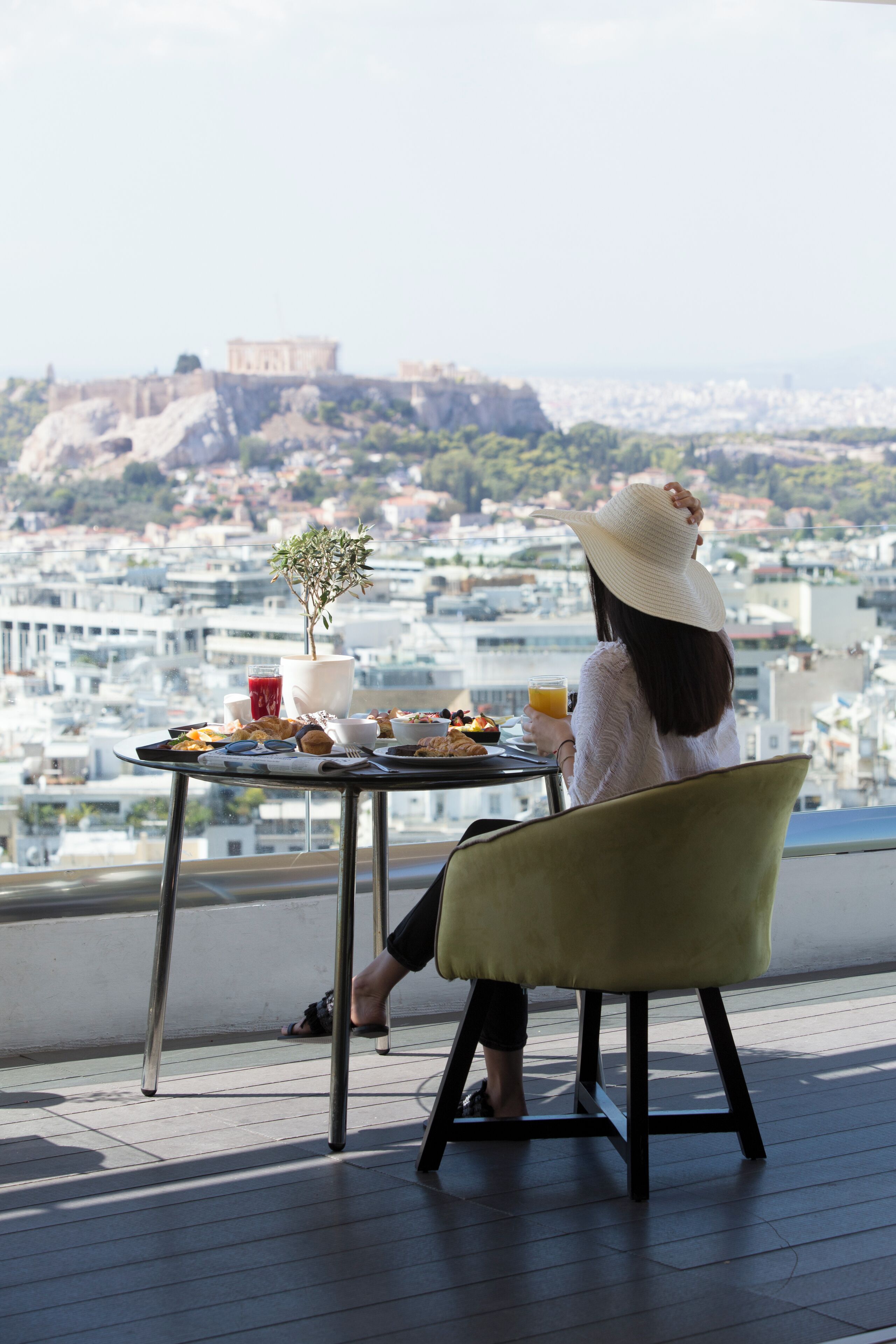 Une femme est assise à une table, avec vue sur la ville, et tient dans ses mains un verre et une assiette de nourriture.