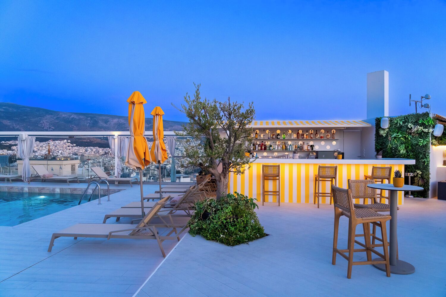Un bar en la azotea con piscina, sillones de descanso y una zona de bar con vistas a la ciudad.