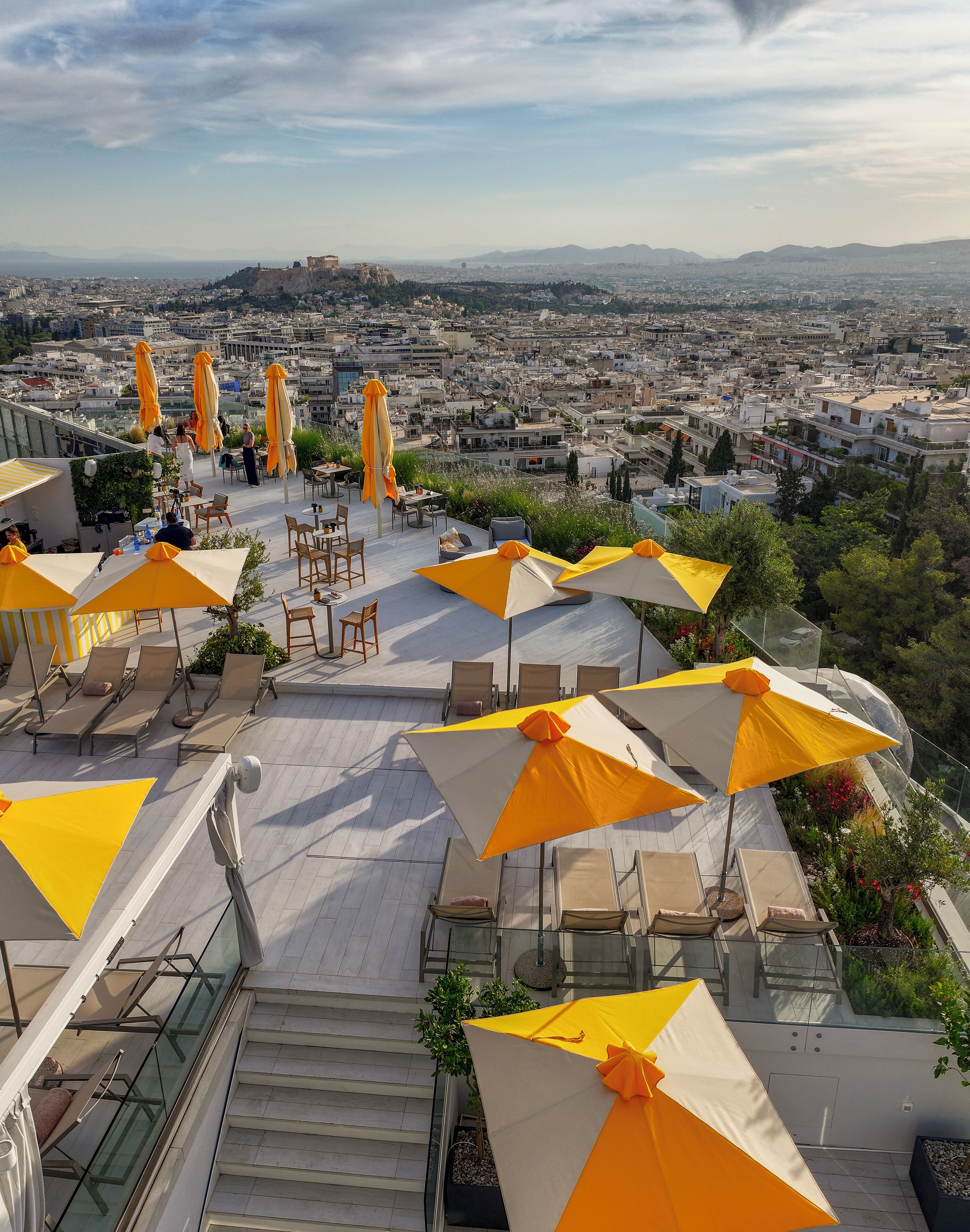 A rooftop terrace with outdoor seating and umbrellas, overlooking a cityscape with buildings and a distant hill.
