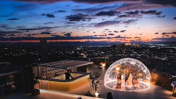A rooftop terrace with a swimming pool, a glass-enclosed lounge area, and a view of the cityscape at dusk.