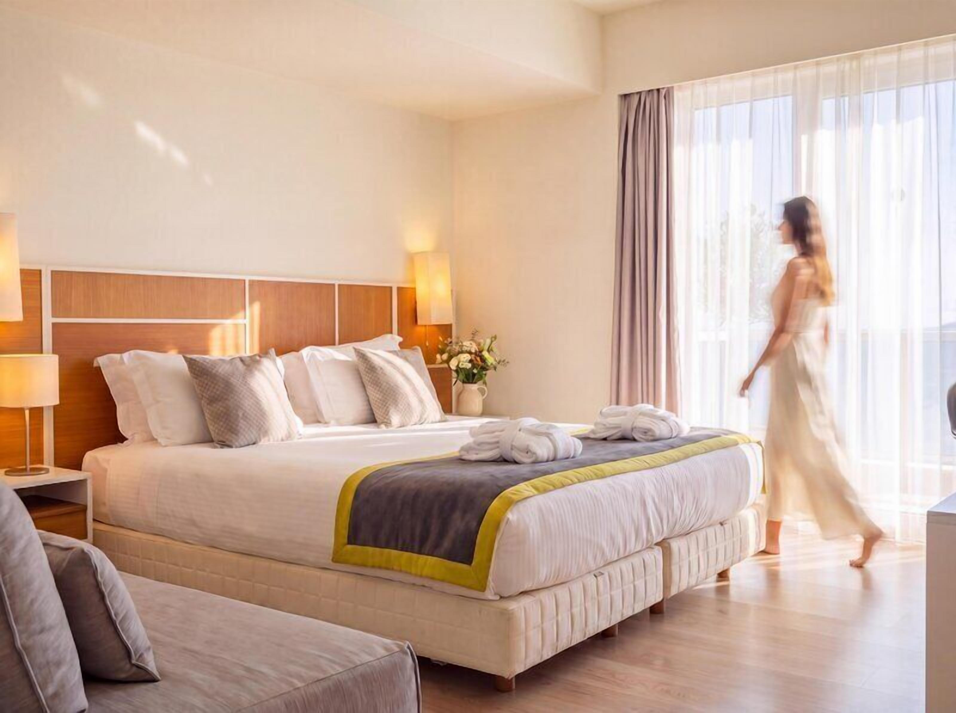 Een hotelkamer met een groot bed, nachtlampjes, een nachtkastje en een raam met gordijnen.