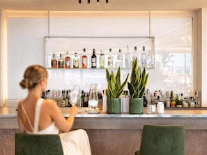 A bar area with a woman seated, a glass of drink, and various bottles on shelves.