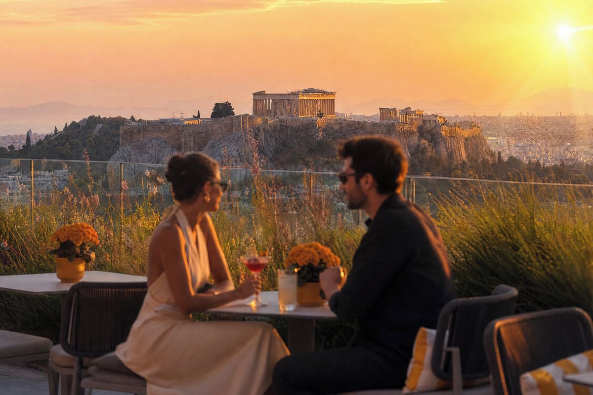 Una pareja cenando en una mesa al aire libre con vistas a ruinas antiguas y un paisaje urbano al fondo.