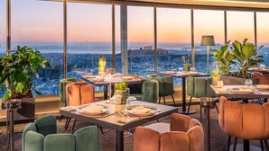 A restaurant with tables set for dining, large windows offering a city view, and potted plants. - St. George Lycabettus Lifestyle Hotel (Athens)