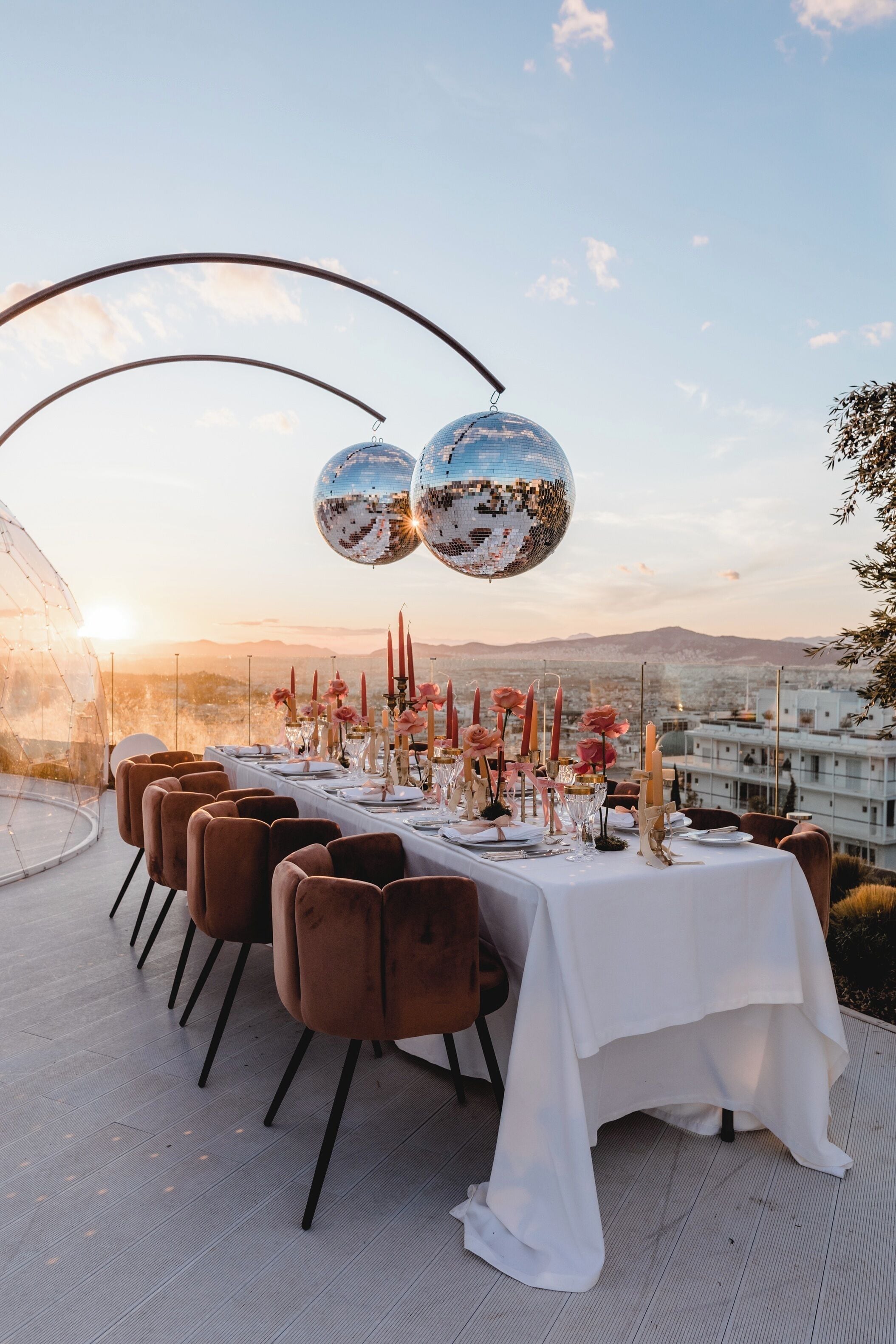 Mesa al aire libre dispuesta para seis personas, con mantel blanco, sillas marrones y elementos decorativos como velas y flores.