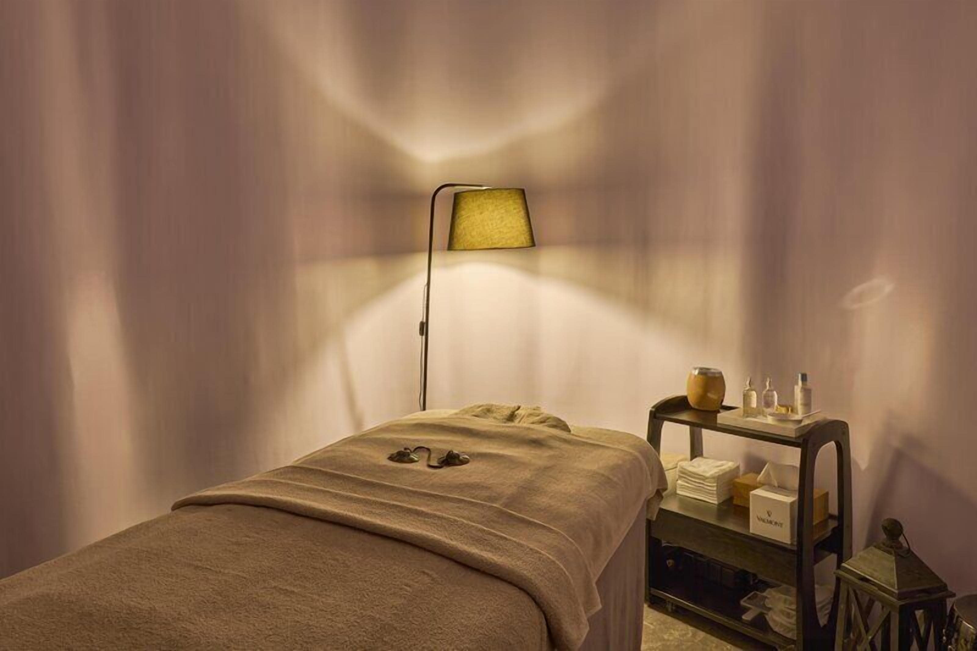 A room with a bed, a bedside table with toiletries, and a lamp.