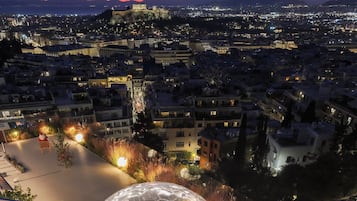 A transparent spherical structure with seating inside, surrounded by a paved area with plants and a view of a cityscape at dusk.