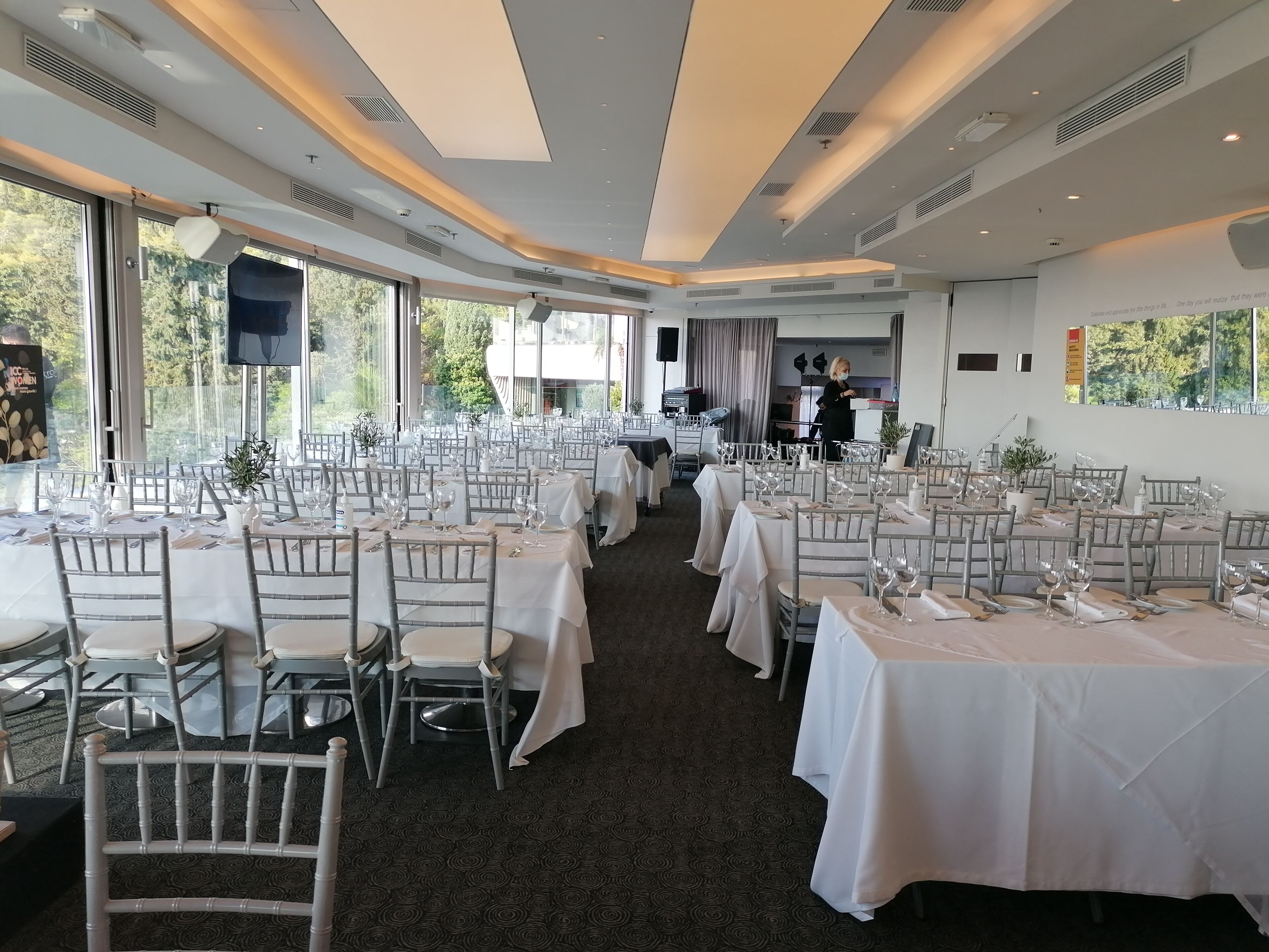 Een ruime eetruimte met meerdere gedekte tafels voor een formeel evenement, grote ramen die natuurlijk licht binnenlaten, en een modern plafondontwerp.