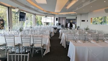 A spacious dining area with multiple tables set for a formal event, large windows offering natural light, and a modern ceiling design.