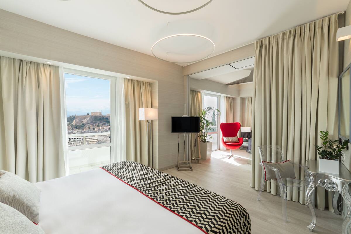 A modern hotel room with a large bed, a TV, a red chair, and a view of a cityscape.