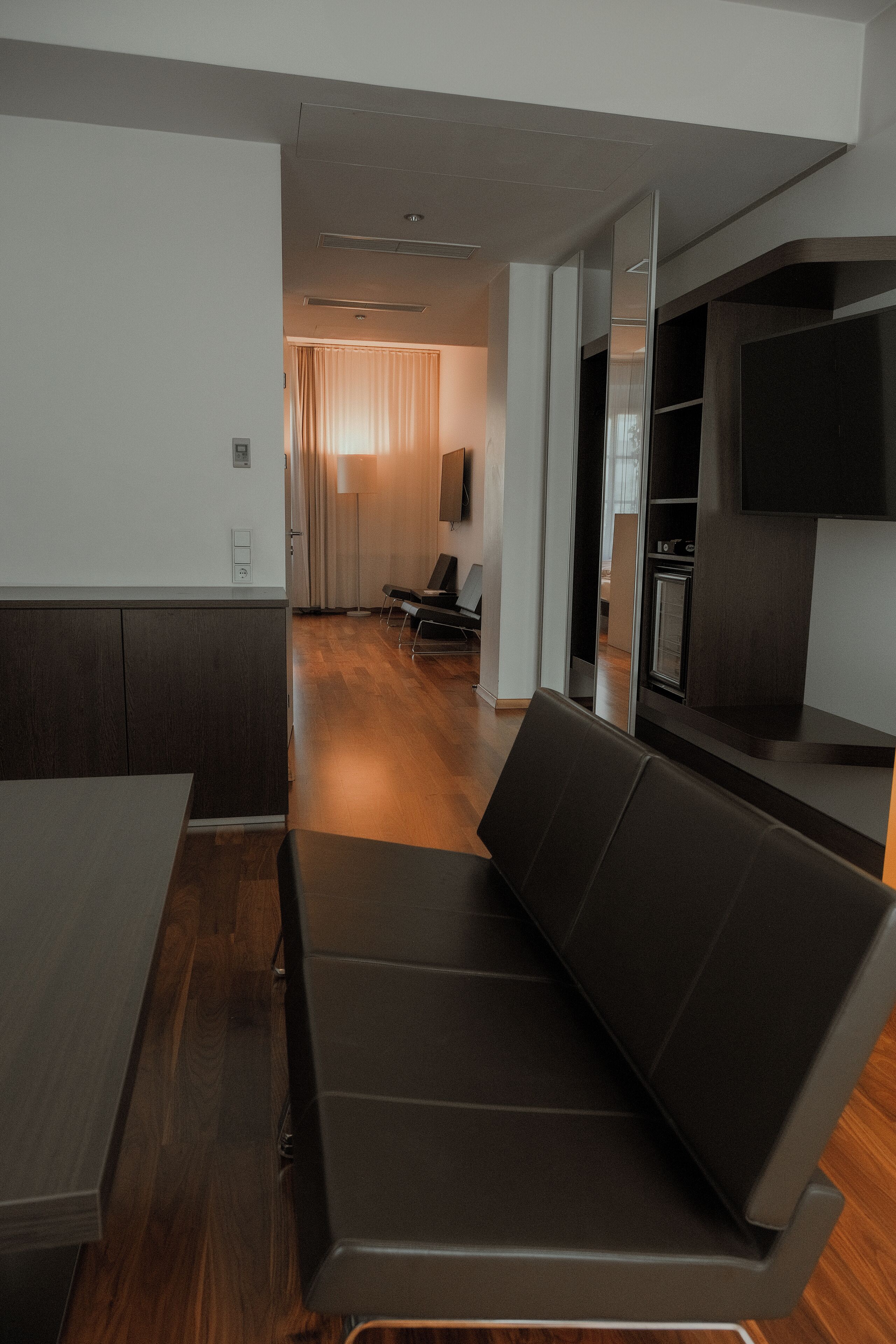 Un interior moderno con suelo de madera, un sofá marrón oscuro, un televisor de pantalla plana y una cocina al fondo.