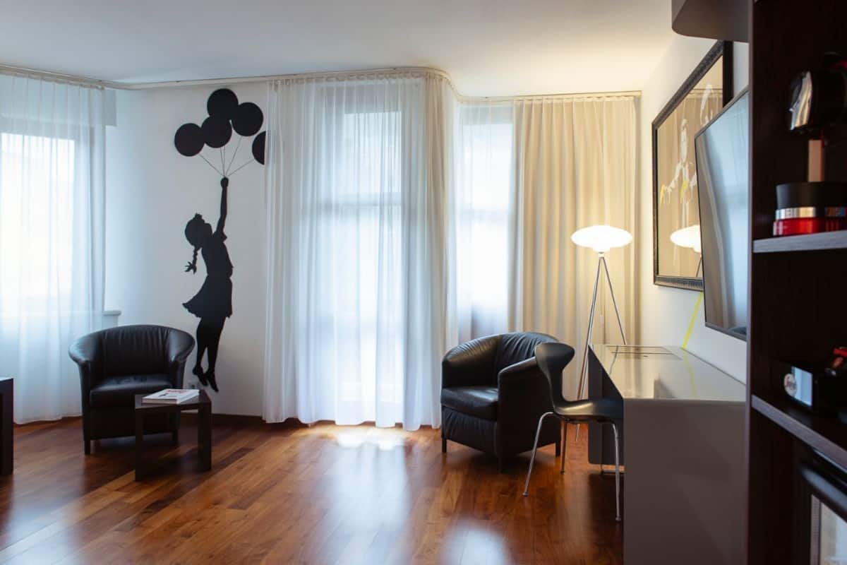 A living room with a wooden floor, two black leather sofas, a glass coffee table, and a large window with sheer curtains.
