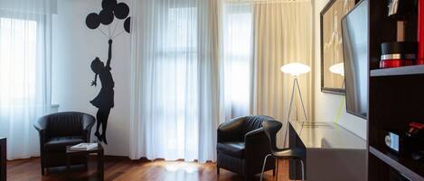 A living room with a wooden floor, two black leather sofas, a glass coffee table, and a large window with sheer curtains.