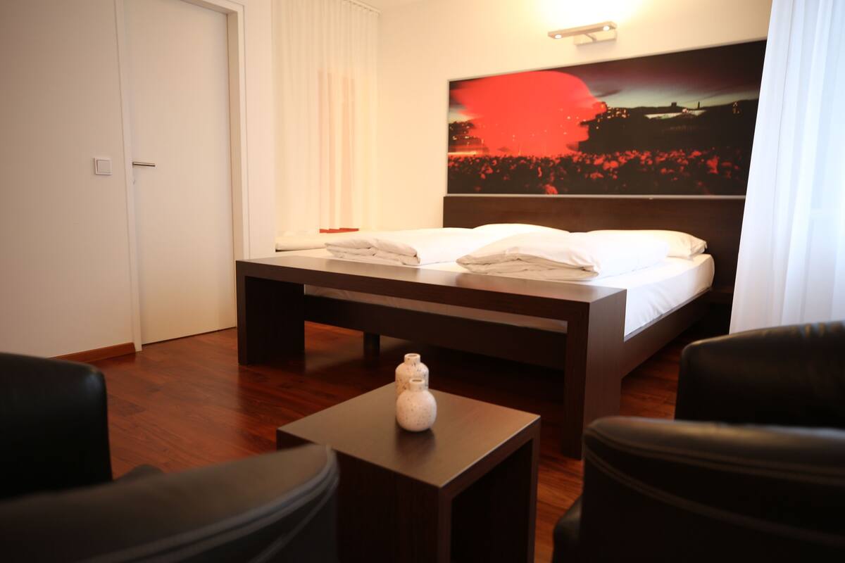 A hotel room with a bed, a bedside table with a vase, a wooden floor, and a large painting on the wall.