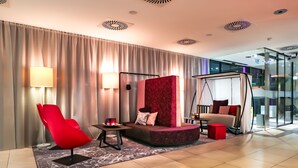 A modern hotel lobby with a red chair, a sofa with a red accent wall, and a glass partition. - arte Hotel Linz (Linz)