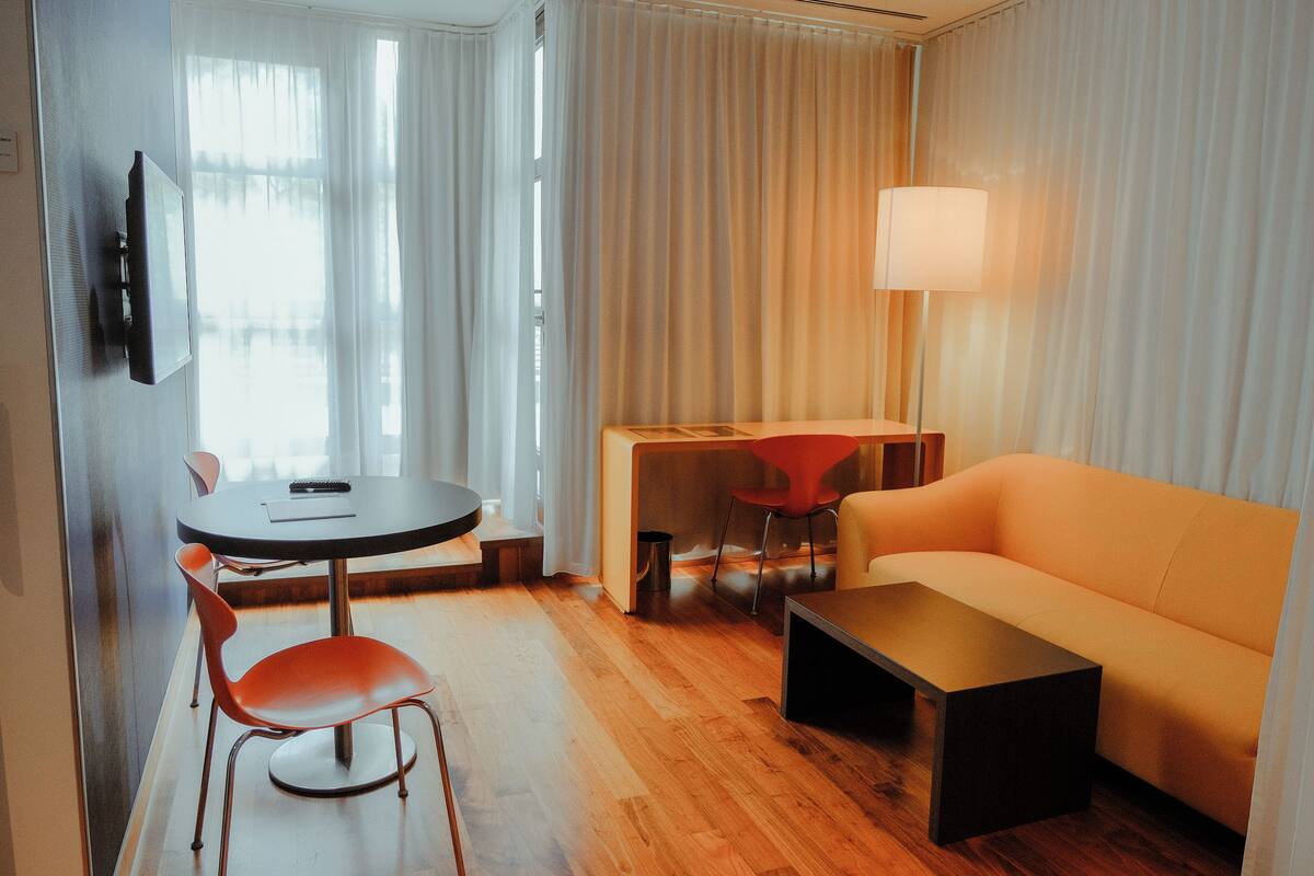 A modern hotel room with a sofa, a dining table with chairs, a TV mounted on the wall, and a lamp.