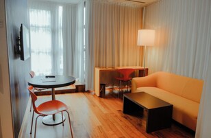 Una habitación de hotel moderna con un sofá, una mesa de comedor con sillas, un televisor montado en la pared y una lámpara.
