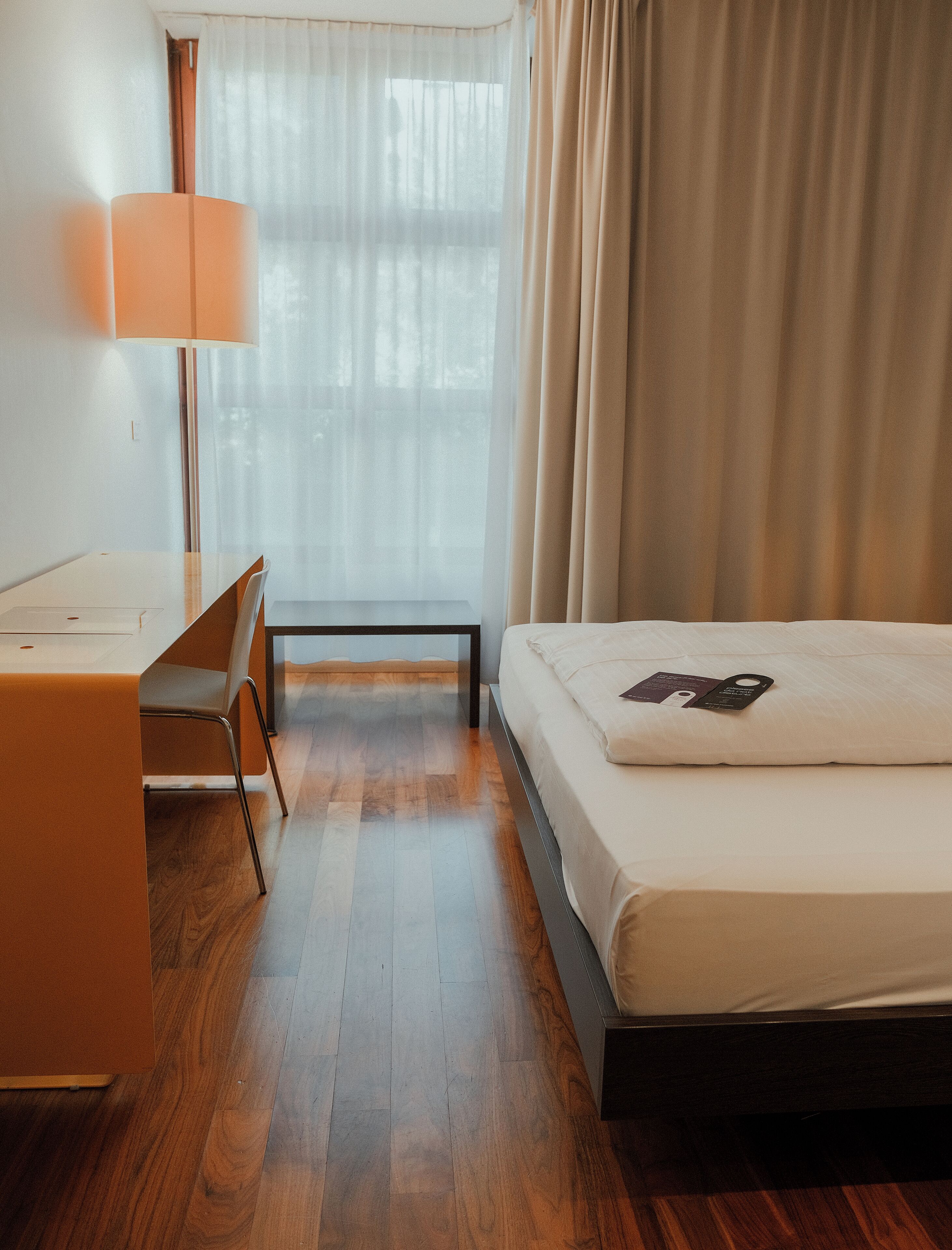 A hotel room with a wooden floor, a desk with a chair, a bed with a pillow, and a window with curtains.